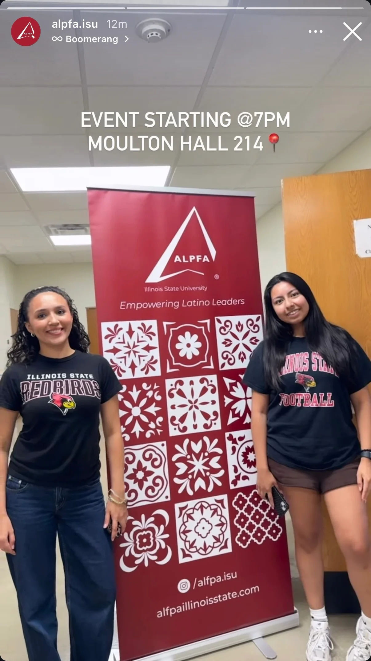 Two members smiling beside the red ALPFA retractable banner, which shows the ALPFA logo, Illinois State University, Empowering Latino Leaders and a grid of white decorative tiles, outside Moulton Hall 214