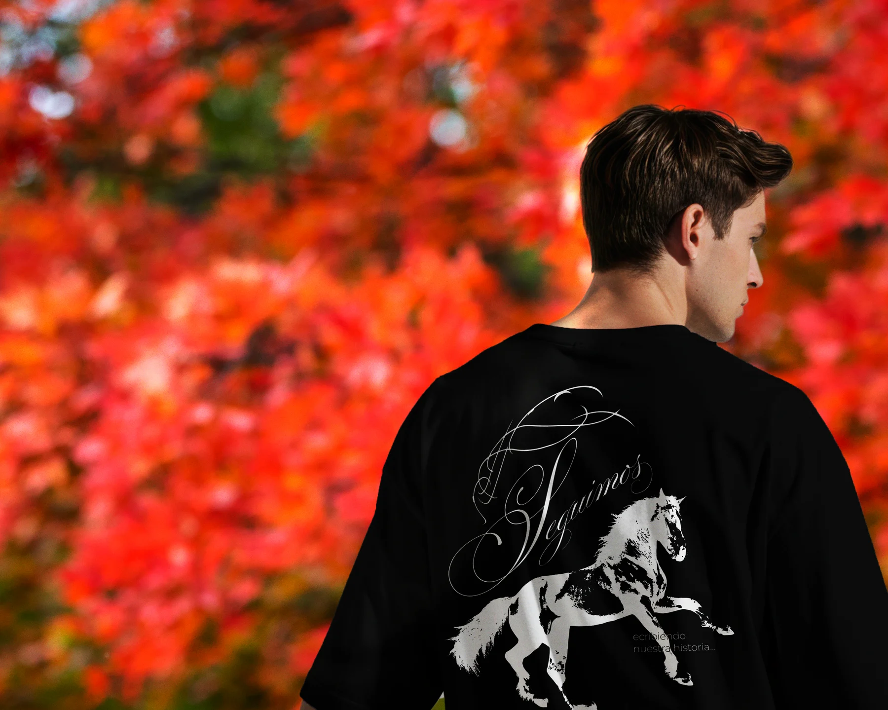 A model seen from behind wearing the black Seguimos shirt, its script lettering and white rearing horse standing out against bright red autumn leaves