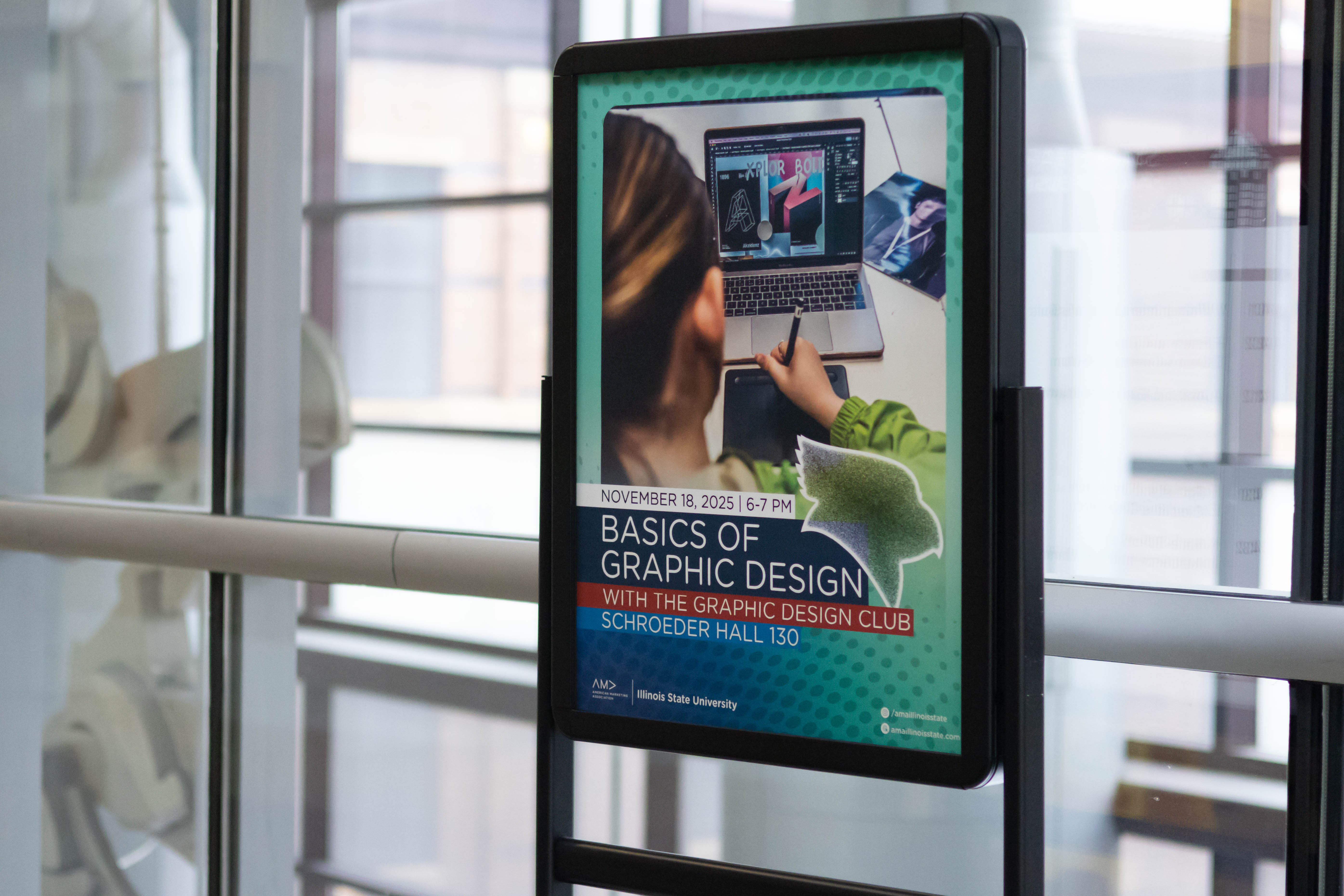 A poster display positioned in front of large glass windows with reflections of the building’s interior. The flyer inside the frame advertises “Basics of Graphic Design” and shows a person using a stylus on a laptop with graphic design software. Soft natural light comes through the windows behind it.