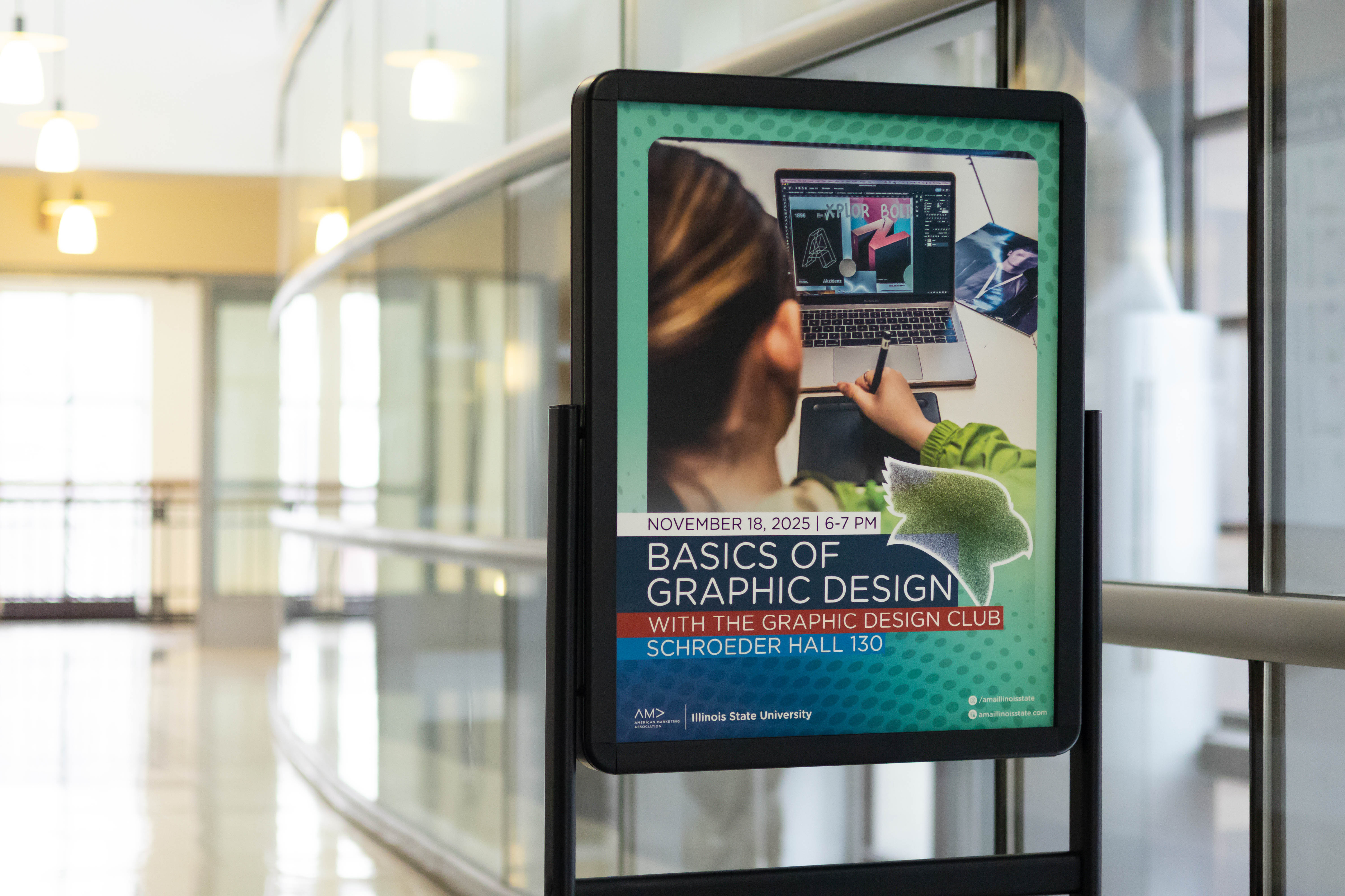 A close-up angle of the same “Basics of Graphic Design” poster placed along a curved glass railing in a bright hallway. Overhead lights hang in the background, and the flyer image of a person working on a laptop is clearly visible within the display frame.