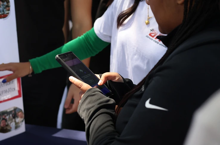 A student scanning a QR code on her phone at an AMA recruitment table, next to a member wearing the AMA Illinois State shirt