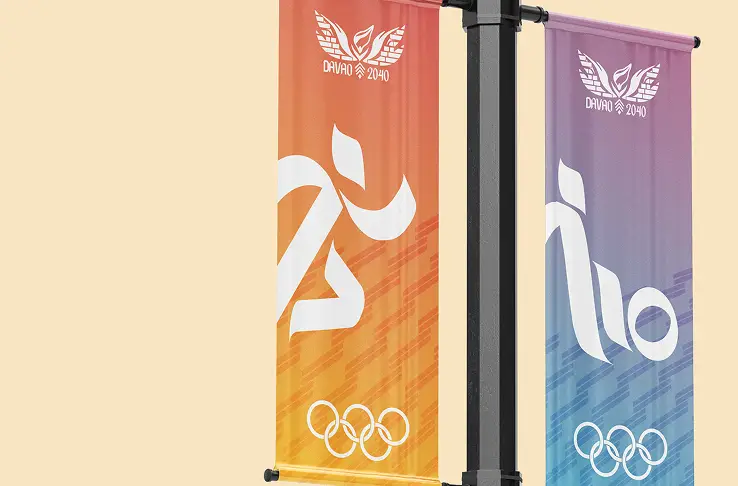 Two vertical street banners hang from a black lamppost, each featuring a colorful gradient background and a white abstract athlete icon. Both banners display the Olympic rings near the bottom and a “Davao 2040” logo with wing-like elements at the top.