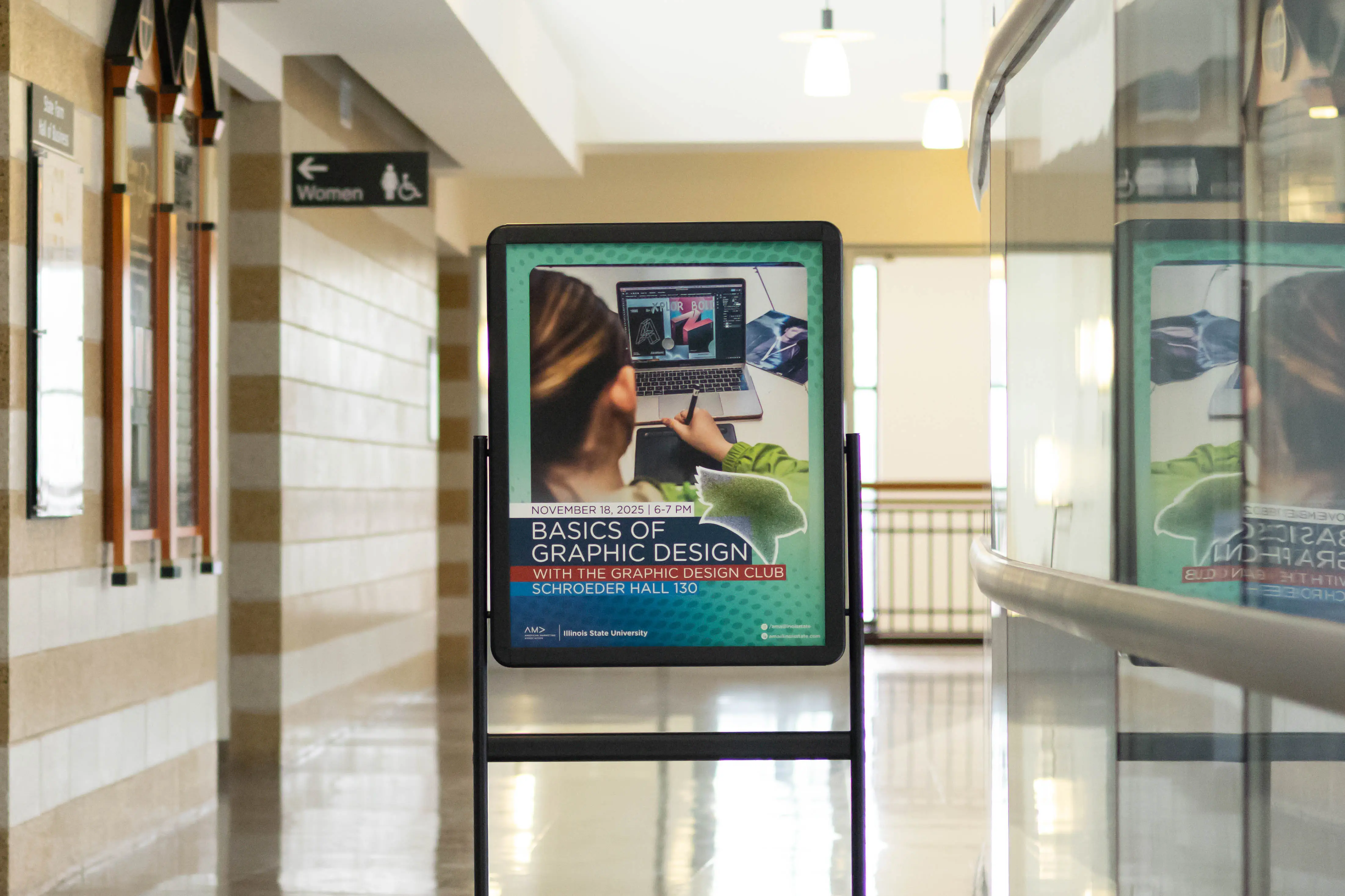 A hallway inside an academic building with beige brick walls and a women’s restroom sign in the background. In the center of the hallway stands a black framed poster display showing a flyer titled “Basics of Graphic Design.” The flyer features a person working on a laptop with design software open and event details for a graphic design workshop.